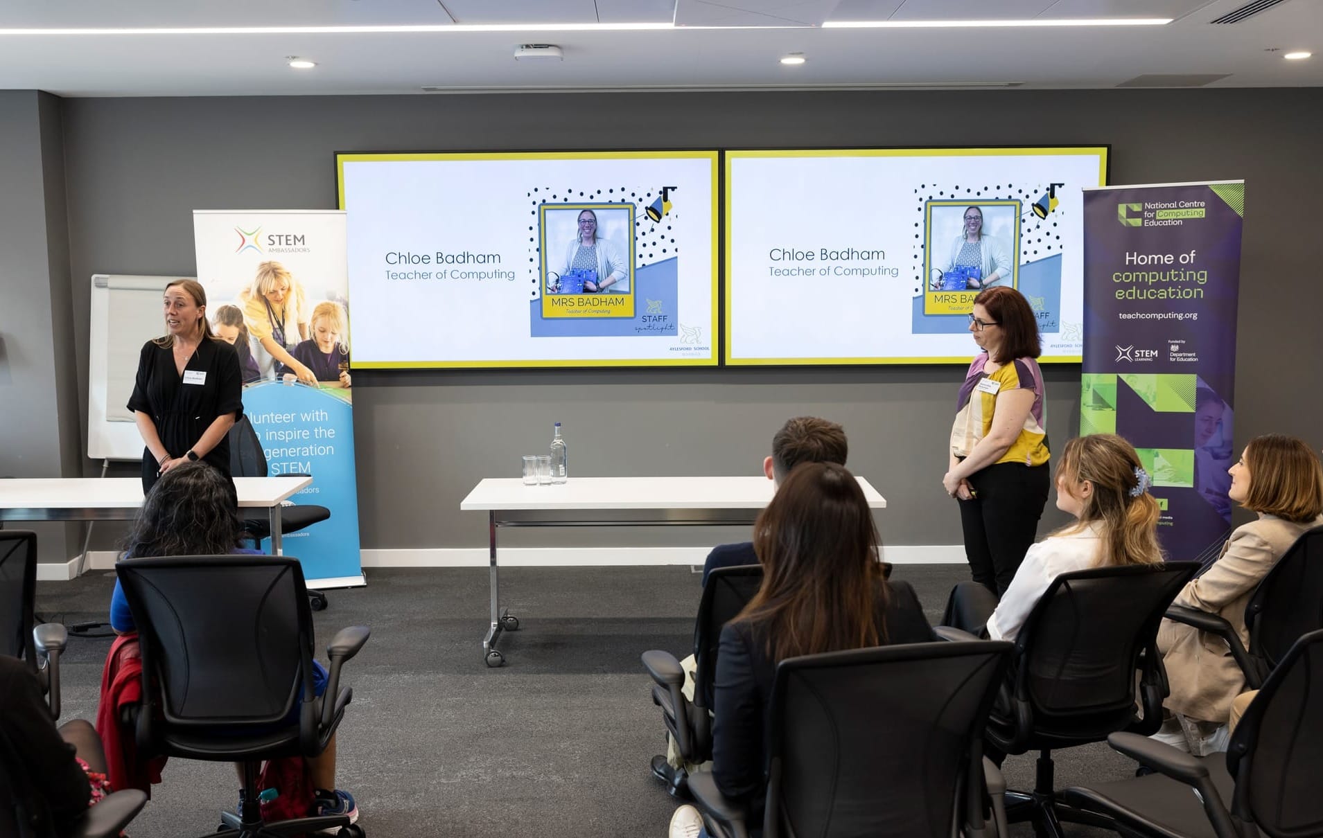 Two presenters in a classroom with screens showing a woman with children using computers.